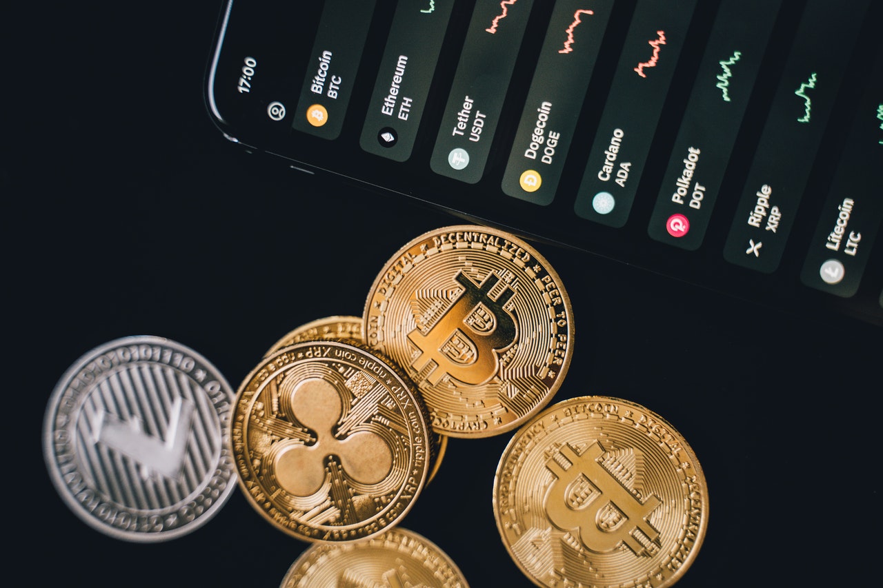 Physical bitcoin and other cryptocoins next to a phone screen displaying a list of digital assets. 