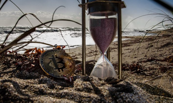 Photo of a bitcoin burried in sand with an hour glass next to it.